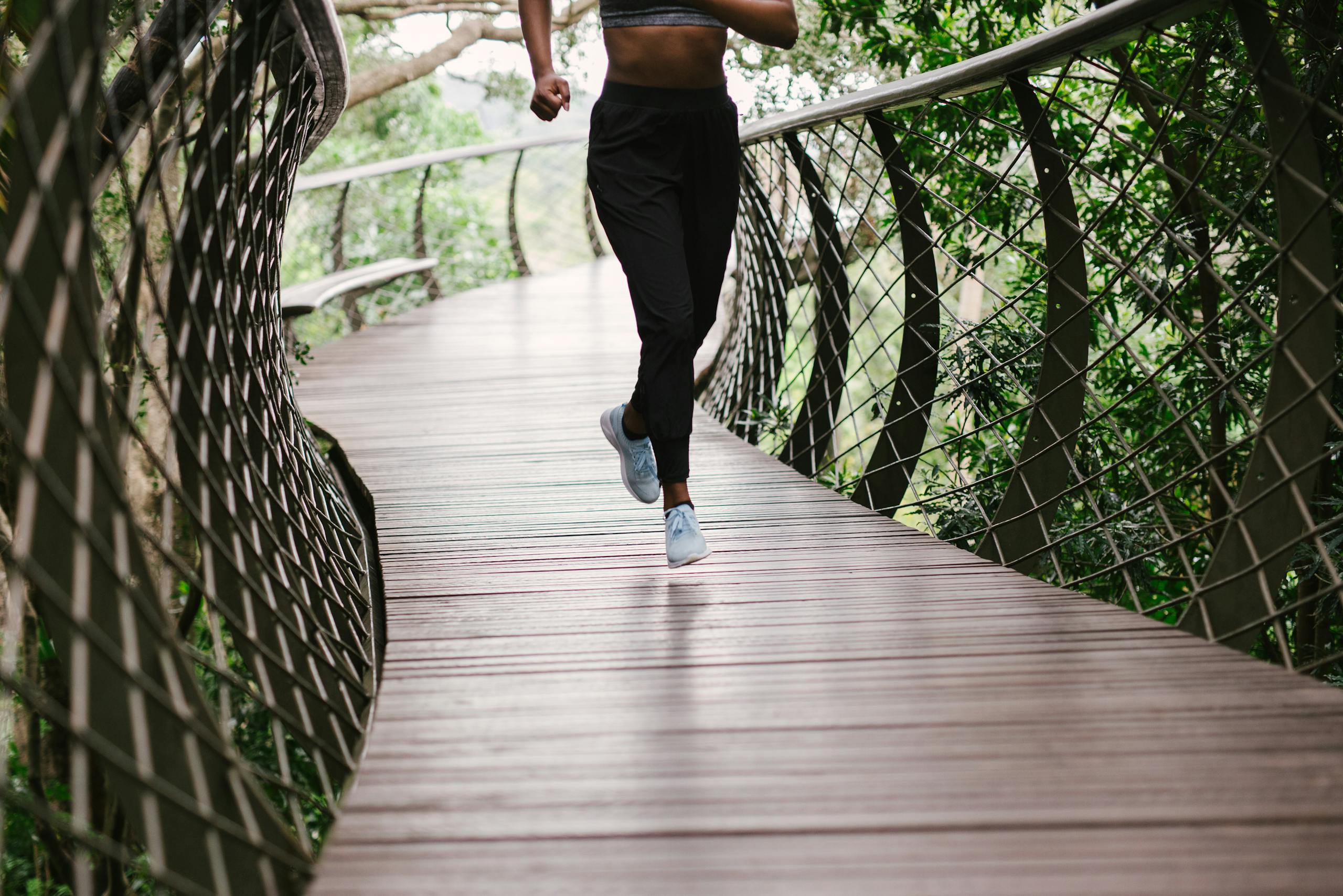 An athletic woman jogging on a nature trail bridge surrounded by greenery.